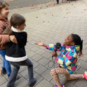 Drei Mädchen machen Quatsch. Eine kniet theatralisch auf dem Boden mit dem Mikrofon und spricht die anderen beiden an, die sich aneinander festklammern und vorbeigehen.