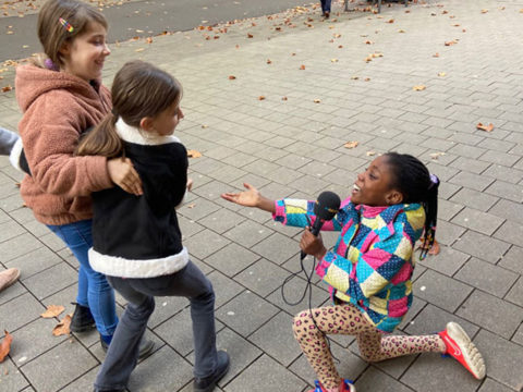 Drei Mädchen machen Quatsch. Eine kniet theatralisch auf dem Boden mit dem Mikrofon und spricht die anderen beiden an, die sich aneinander festklammern und vorbeigehen.