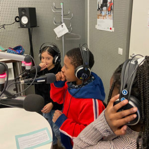 Drei Kinder, ein Junge und zwei Mädchen, stehen dicht vor drei Mikrofonen in einem Radiostudio. Sie tragen Kopfhörer und der Junge hält seine Faust zwischen Mund und Mikrofon.