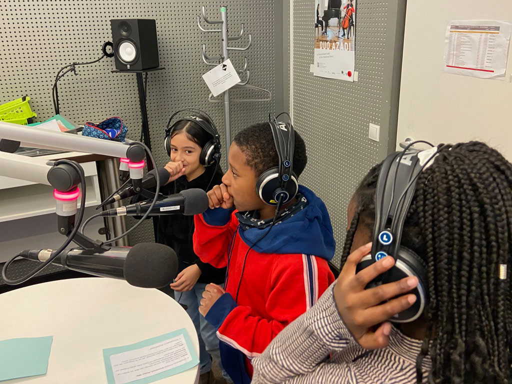 Drei Kinder, ein Junge und zwei Mädchen, stehen dicht vor drei Mikrofonen in einem Radiostudio. Sie tragen Kopfhörer und der Junge hält seine Faust zwischen Mund und Mikrofon.