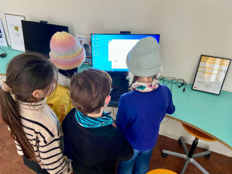 Vier Kinder stehen vor einem Computer und gucken gespannt auf den Monitor.