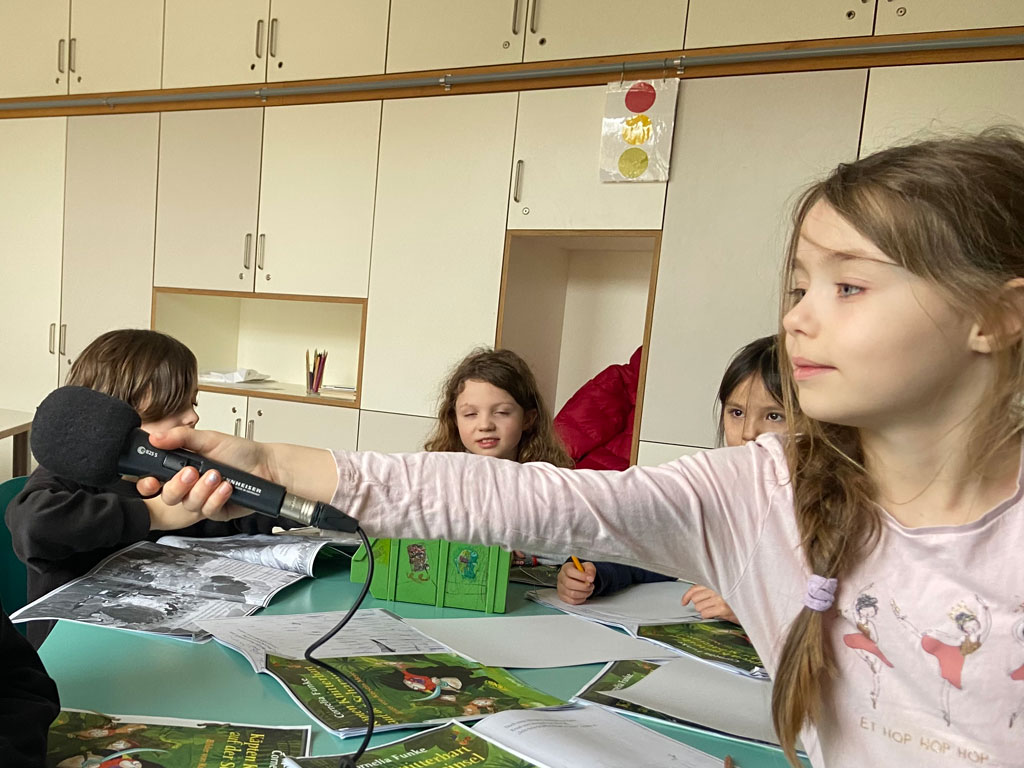 Ein Mädchen hält ein Mikrofon mit dem ausgestreckten Arm. Drei weitere Kinder sitzen im Hintergrund.