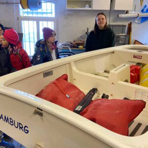 Drei Kinder stehen neben einem kleinen Segelboot in einem Raum. Eine Frau erklärt ihnen etwas.