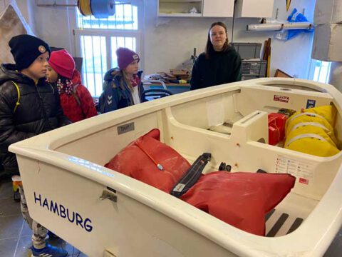 Drei Kinder stehen neben einem kleinen Segelboot in einem Raum. Eine Frau erklärt ihnen etwas.