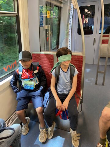 Zwei Kinder sitzen in der U-Bahn. Eines hat die Augen verbunden.