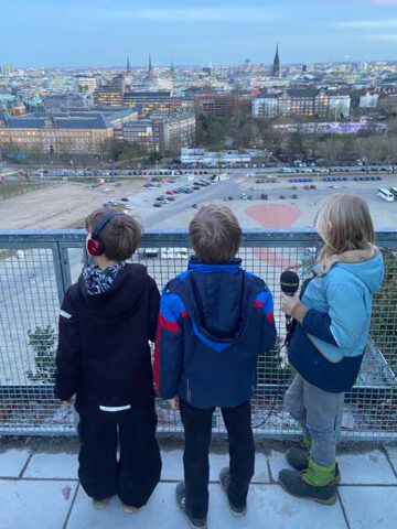 Drei Kinder mit Kopfhörern und Mikrofon von hinten stehen auf einer hohen Plattform und blicken über die Stadt Hamburg