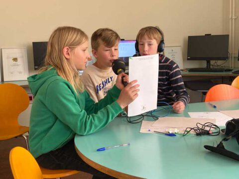Drei Kinder mit Kopfhörern und Mikrofon in einem Computer-Raum lesen von einem Blatt ab