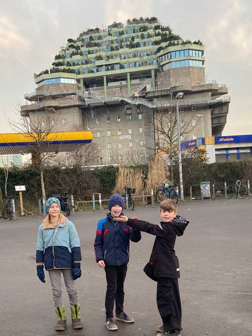 Drei Kinder stehen vor einem hohen Bunker-Gebäude, auf dem Bäume wachsen