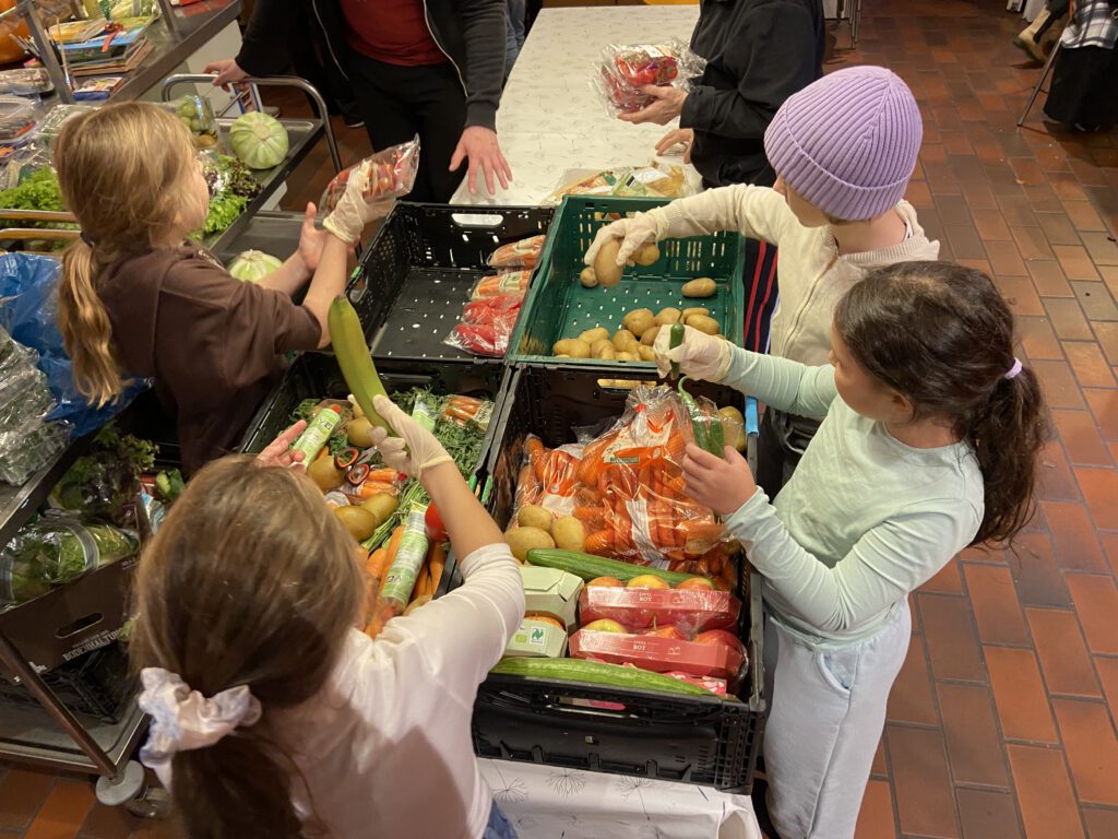 Auf Tischen stehen grüne Kisten mit Gemüse. Drumherum stehen Kinder und helfen beim Sortieren.