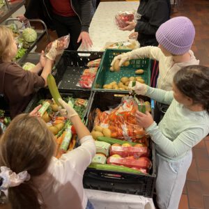 Auf Tischen stehen grüne Kisten mit Gemüse. Drumherum stehen Kinder und helfen beim Sortieren.