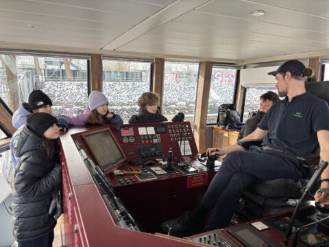 Mehrere Kinderreporter stehen um das Cockpit und den Kapitän der HADAG Fähre herum