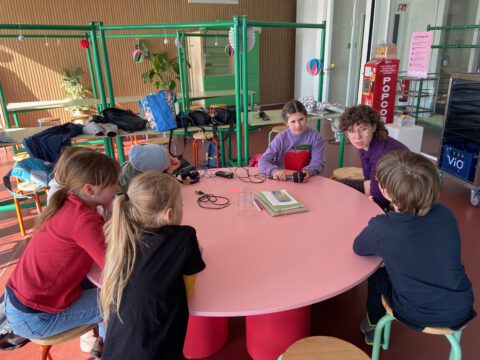 Kinderreporter:innen sitzen um einen runden Tisch und interviewen eine Mitarbeiterin