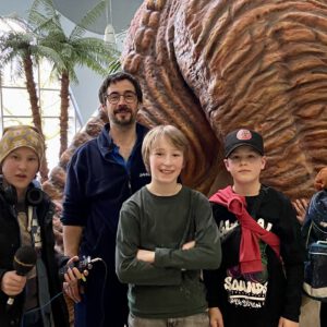 Vier Kinderreporter stehen im Hallenbad mit einem Bademeister vor einem großen Dinosaurier