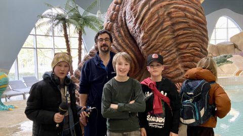 Vier Kinderreporter stehen im Hallenbad mit einem Bademeister vor einem großen Dinosaurier