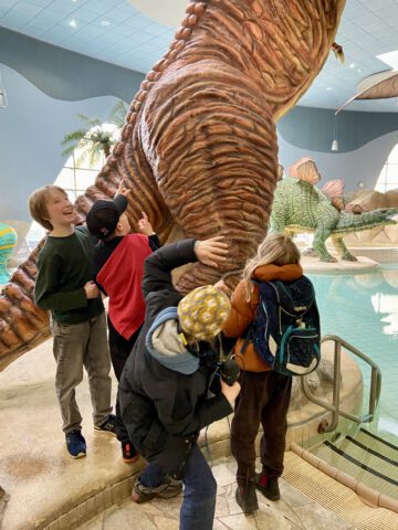 Vier Kinderreporter stehen nah am großen Dino und berühren ihn mit den Händen