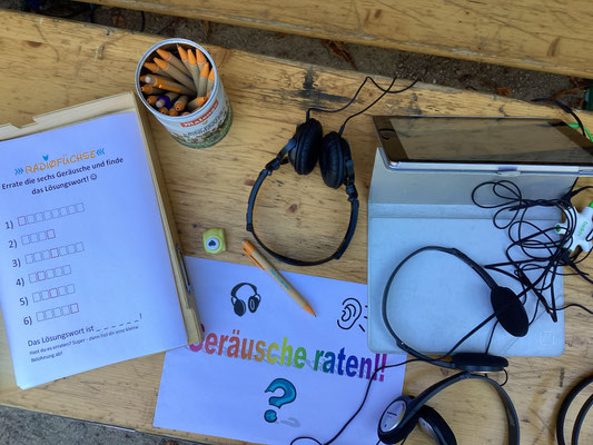 Ein Tisch von oben fotografiert. Auf dem Tisch liegt ein Zettel, auf dem "Geräuscherätsel" steht. Daneben liegen Kopfhörer, Stifte und ein Tablet.