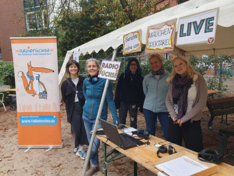 Fünf erwachsene Frauen stehen hinter einem Gartentisch am Stand der Radiofüchse, der mit vieen Schildern dekoriert ist. Auf dem Tisch steht ein Laptop.